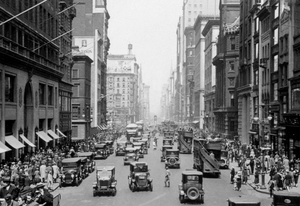 Historijska slika užurbane gradske ulice, ispunjene pješacima i automobilima.